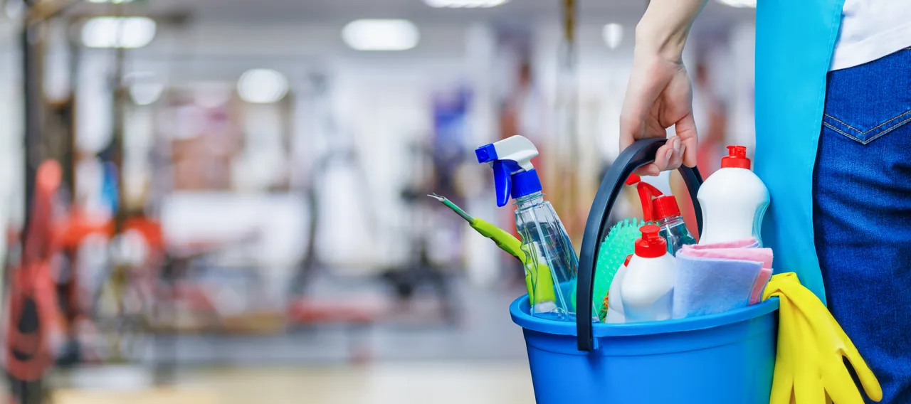 Mano sosteniendo un balde azul con atomizadores, cepillos, paños y guantes para limpiar casas y oficinas en Orlando