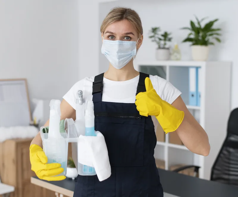 Cleaning professional with a mask and yellow gloves holding cleaning supplies and giving a thumbs up in an office in Orlando